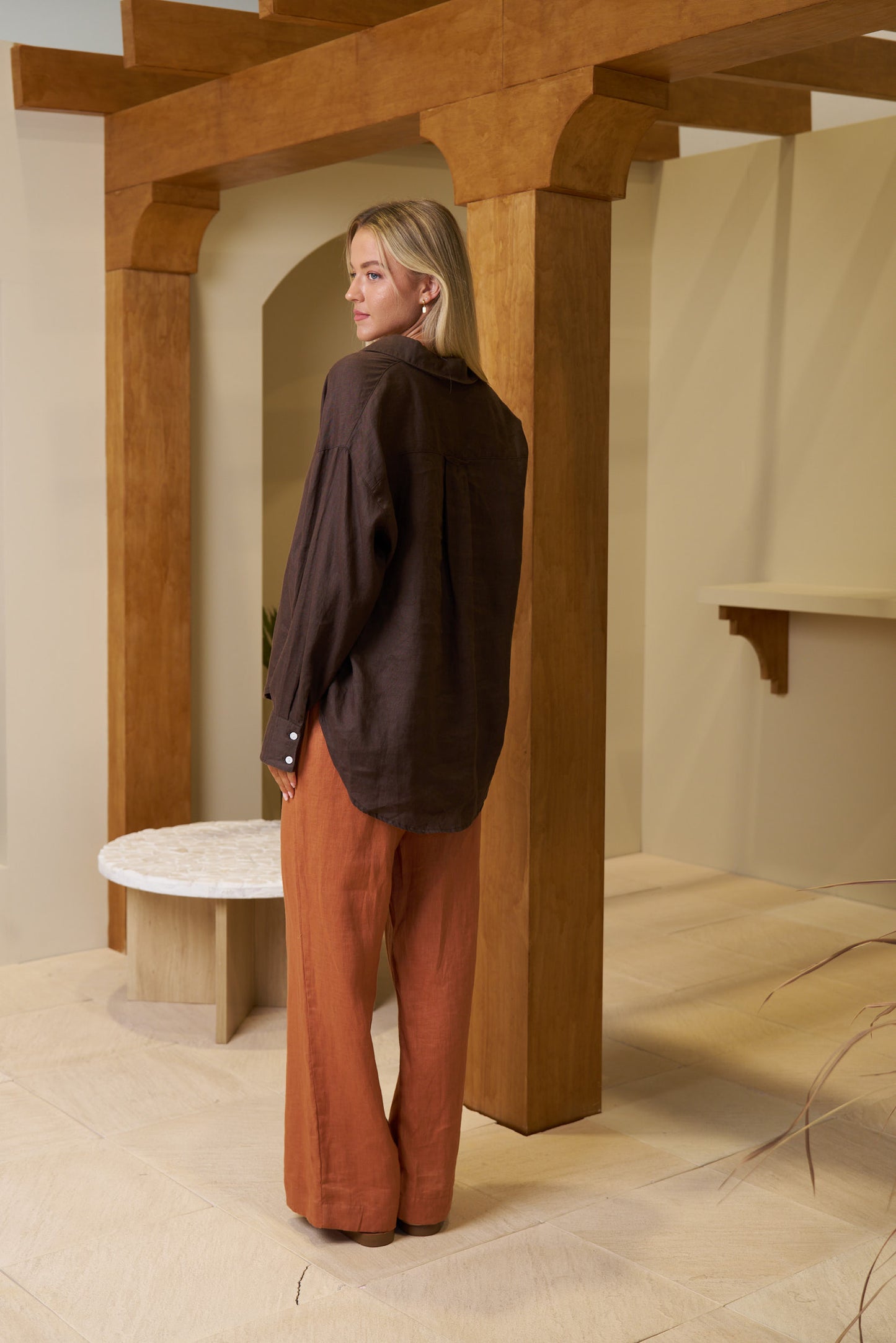 Person wearing a dark sweater and rust-colored pants standing in a room with wooden beams and a white table.